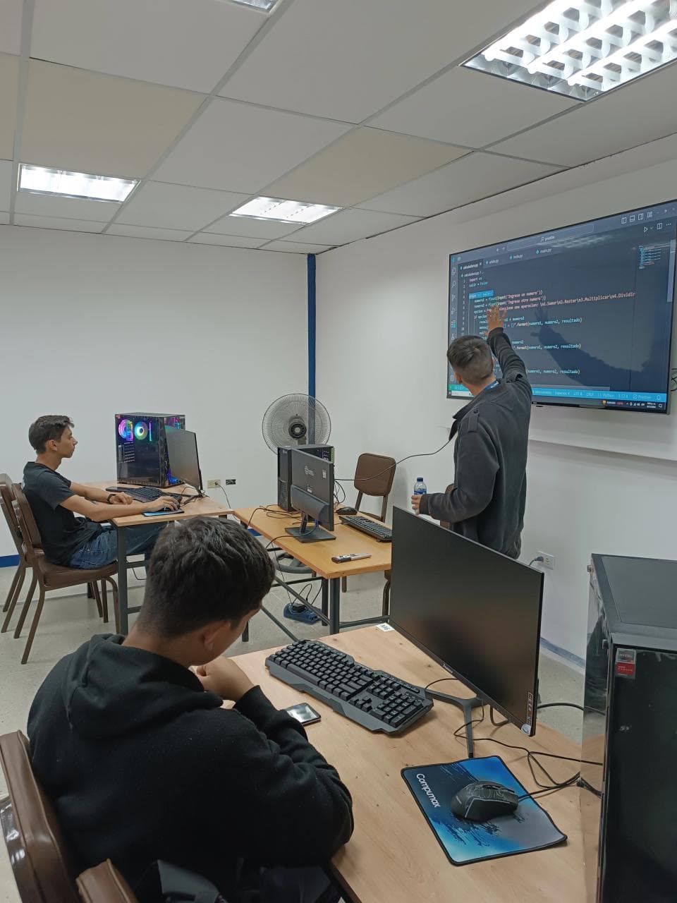 A computer lab: I am at the big screen explaining a Python exercise while two students follow along from their desks.