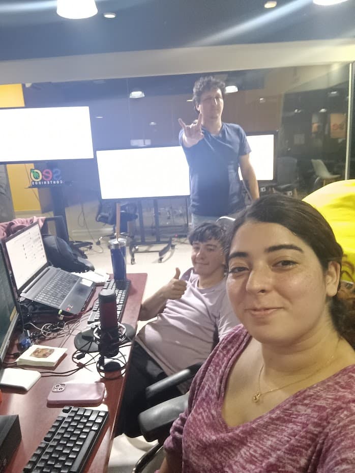 Three of us at the same office, mid-shift, with monitors and a microphone on the desks and the company logo on the wall behind.
