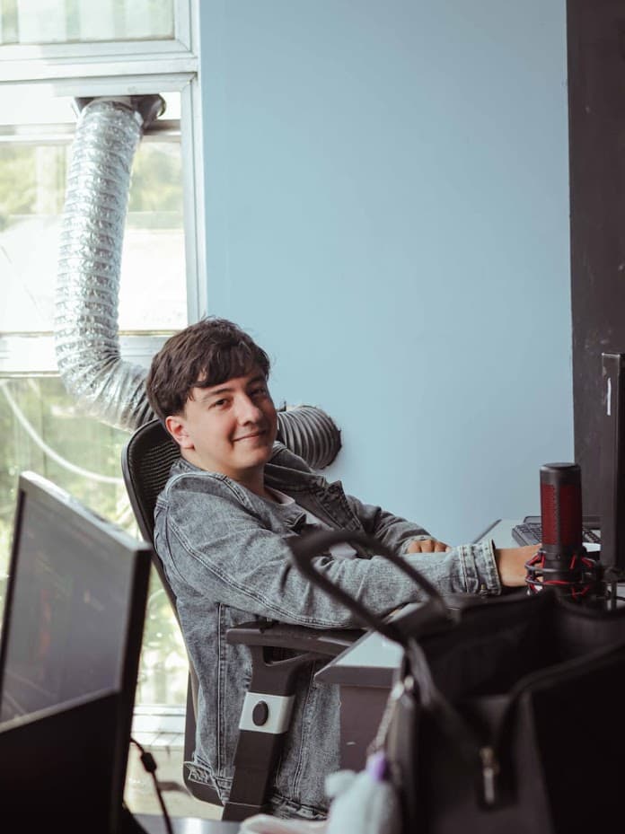 Me at my desk in a denim jacket, turned away from two monitors and a microphone, next to a tall window.